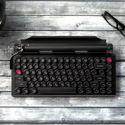 Qwerkywriter typewriter keyboard best in class premium all aluminum body in anniversary dark chrome top angle on grey wood grain background with a pair of glasses