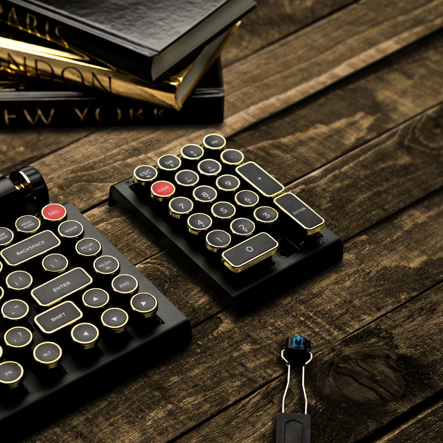 Qwerkywriter typewriter keyboard numkey numpad best in class premium all aluminum body in black gold close view on brown wood grain with hot swap tool cherry mx blue switch and books